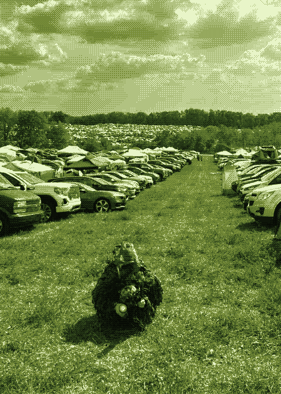 a ghillie-suited figure resting motionless in the grass of a festival campground, rows of parked cars and tents behind it