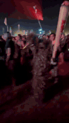 a figure in a ghillie suit moving through a dense festival crowd at night under red and blue lights