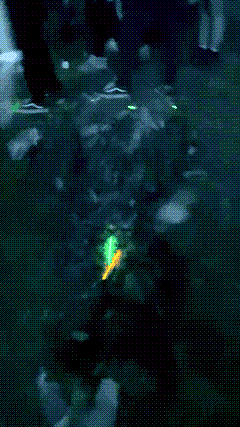a figure in a ghillie suit seated at night, arms raised mid-gesture, surrounded by dropped glowsticks